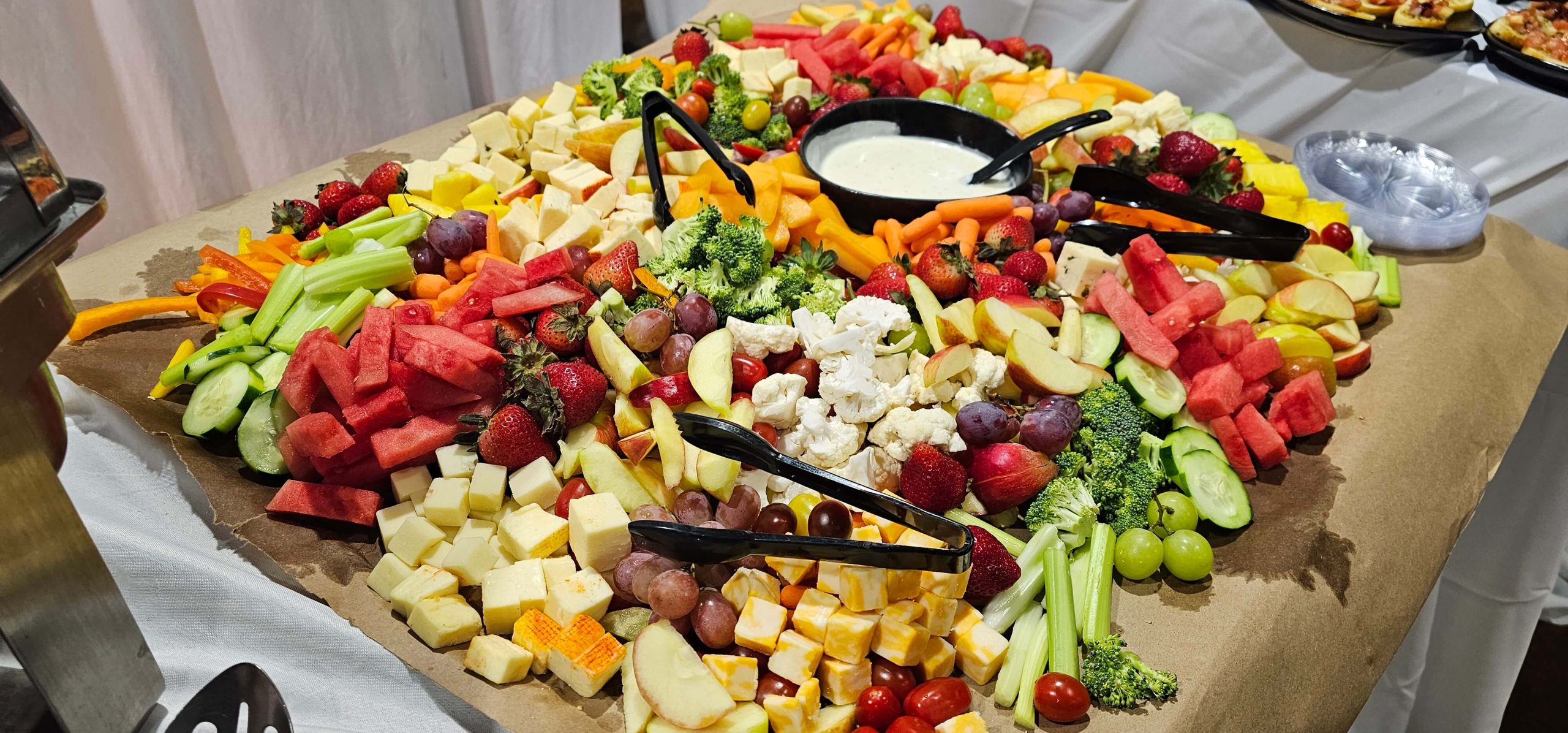 portion of a table overflowing with many fruits and vegetables and cheeses with dip